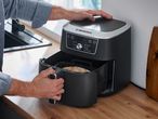 A person operating a black air fryer with a digital display showing cooking modes like REHEAT, ROAST, BAKE, DEHYDRATE, AIR FRY, PROVE, and ULTRA CRISP.