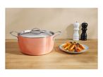 Copper-colored cooking pot with lid, plate of pasta, and salt and pepper grinders on a wooden surface.