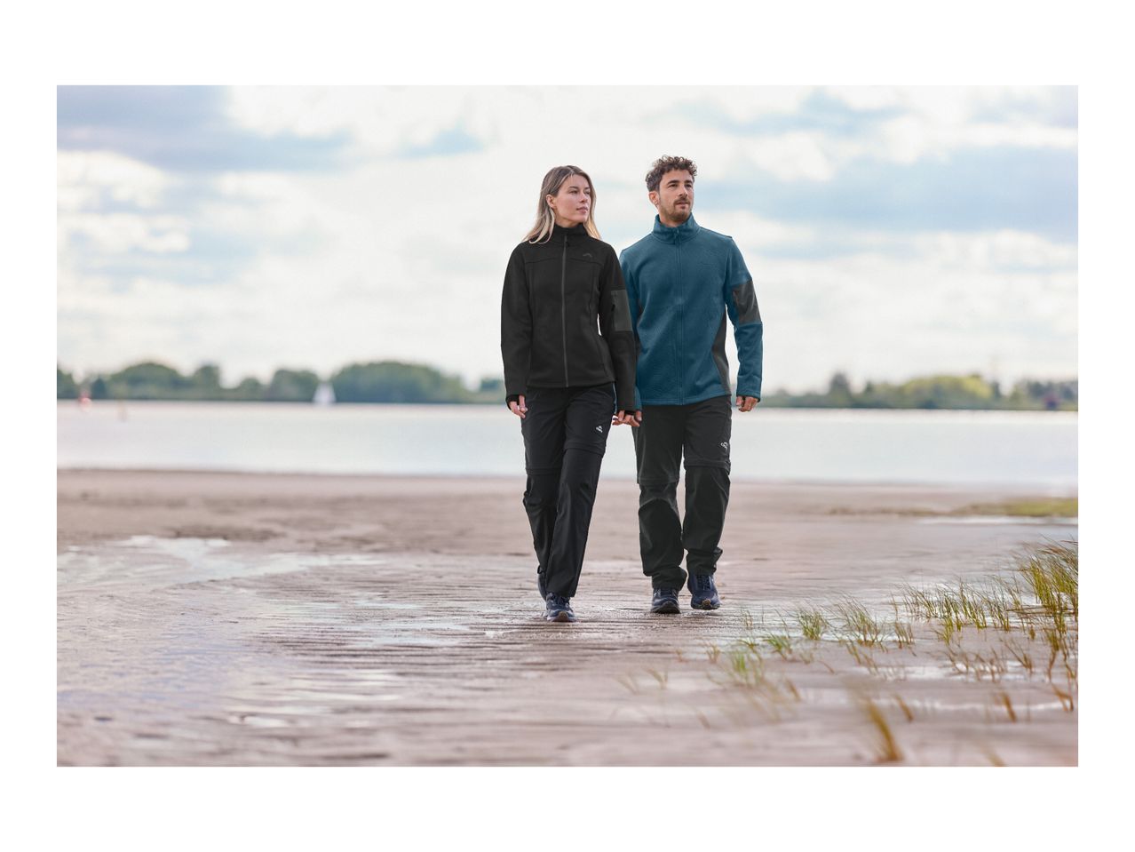 Man and woman in outdoor jackets and convertible trousers walking on a beach.