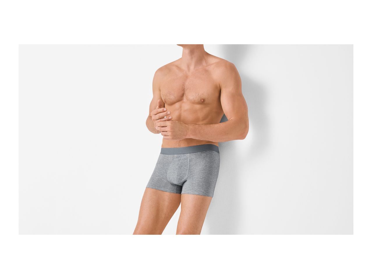 Man in grey boxer briefs leaning against a white wall.