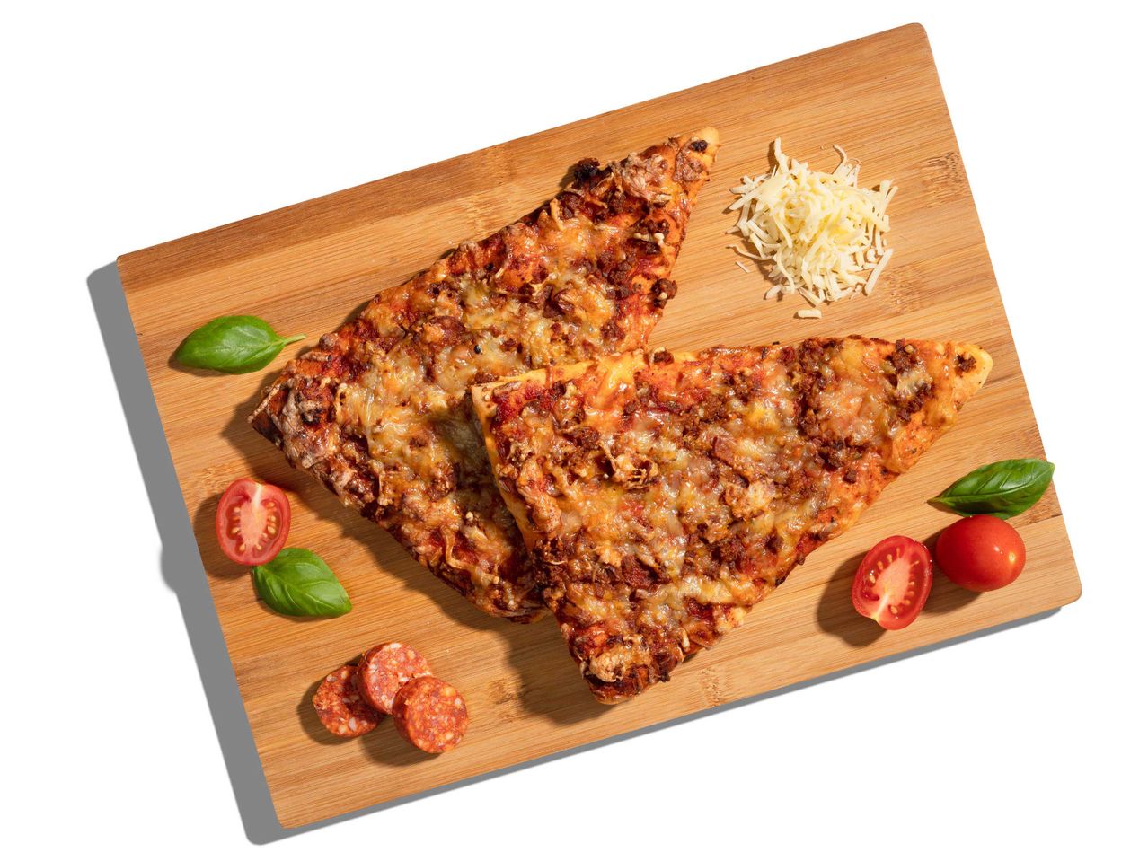 Két szelet pizza egy vágódeszkán, paradicsommal, bazsalikommal, reszelt sajttal és szalámival.