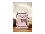 Pink espresso machine with a temperature gauge, two cups, and coffee beans on a wooden counter.