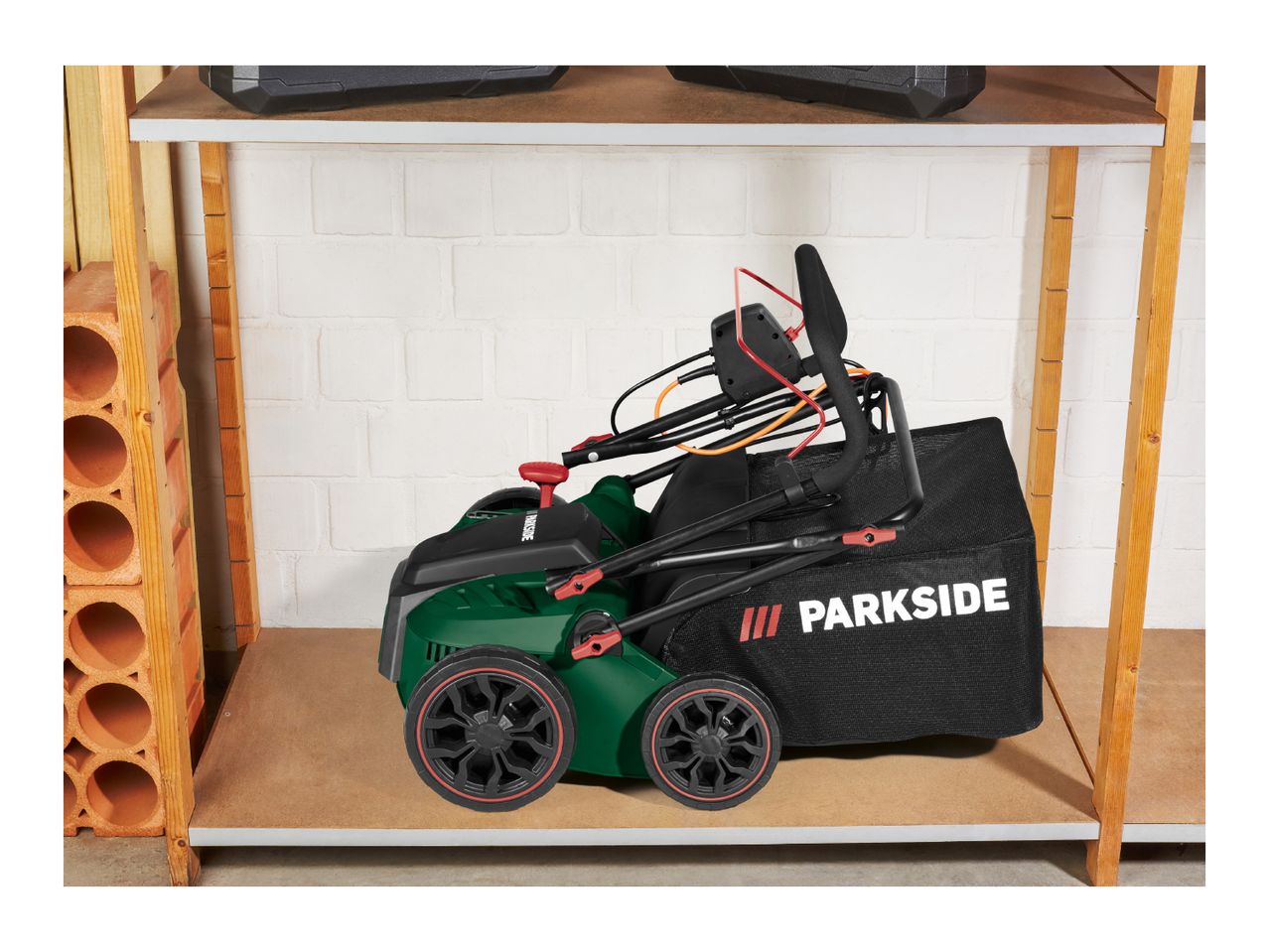 Green lawnmower with a black grass collection bag on a wooden shelf in a storage area.