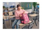A girl in a classroom, sitting on a desk and opening her pink floral backpack.