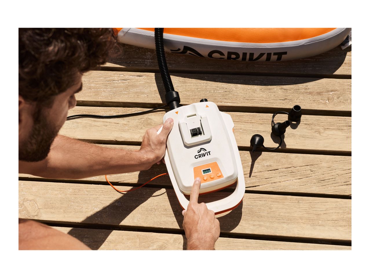 A person operating an electric pump to inflate an orange and white inflatable paddleboard on a wooden deck.