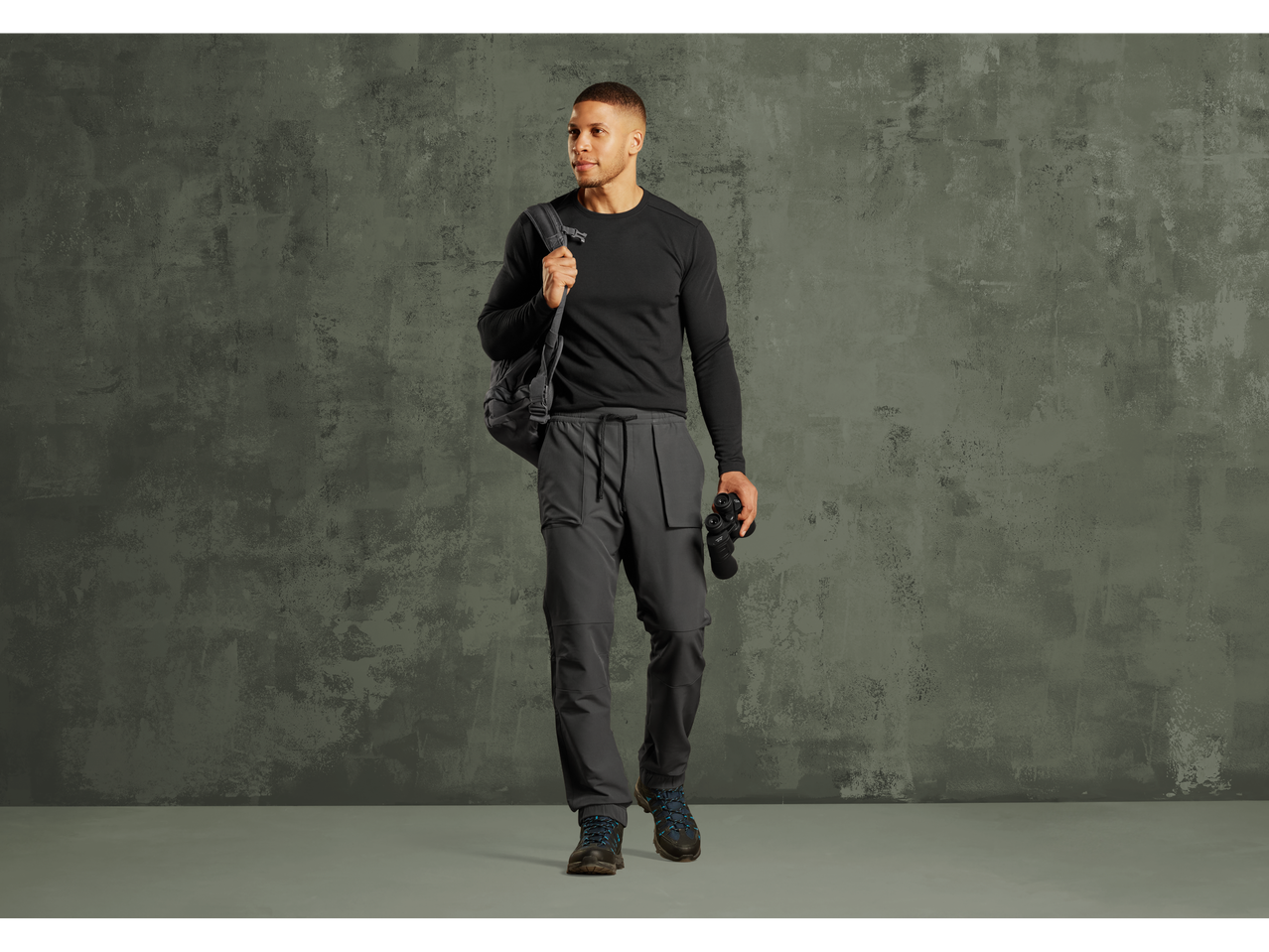 Man in black long-sleeve shirt, gray cargo pants, and hiking boots, holding binoculars.