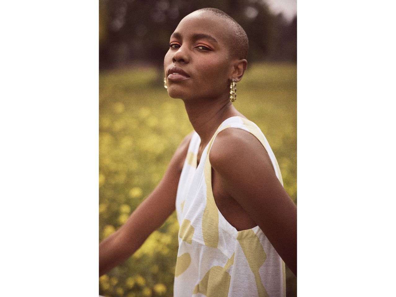 A woman with a shaved head wears a sleeveless, abstract-print dress in a field of yellow flowers.