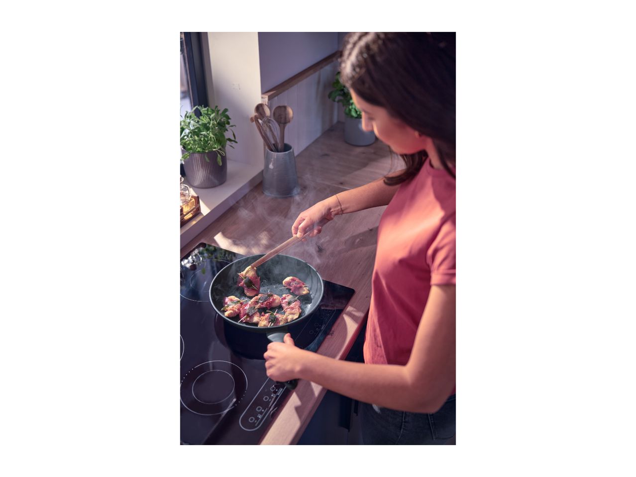 A person cooking meat wrapped in bacon and sage in a frying pan on an induction hob.