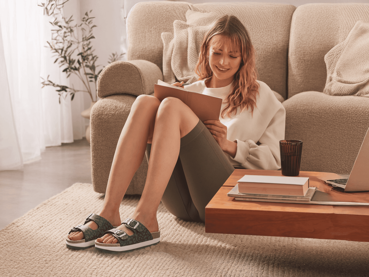 Eine junge Frau sitzt auf einem Sofa und trägt BIRKENSTOCK-Sandalen, während sie in einem Notizbuch schreibt.