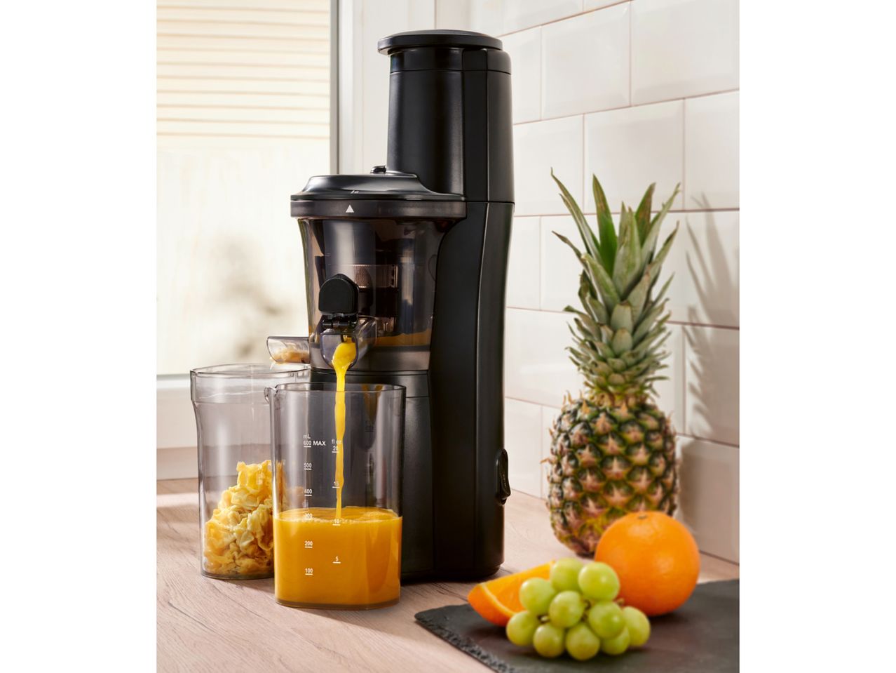 Black slow juicer making orange juice, with pineapple, orange, and grapes nearby.