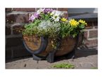 Half-barrel planter with colorful flowers and green plants on a paved surface.
