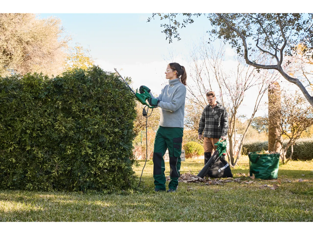 A woman uses a Parkside hedge trimmer while a man uses a leaf blower in a garden.
