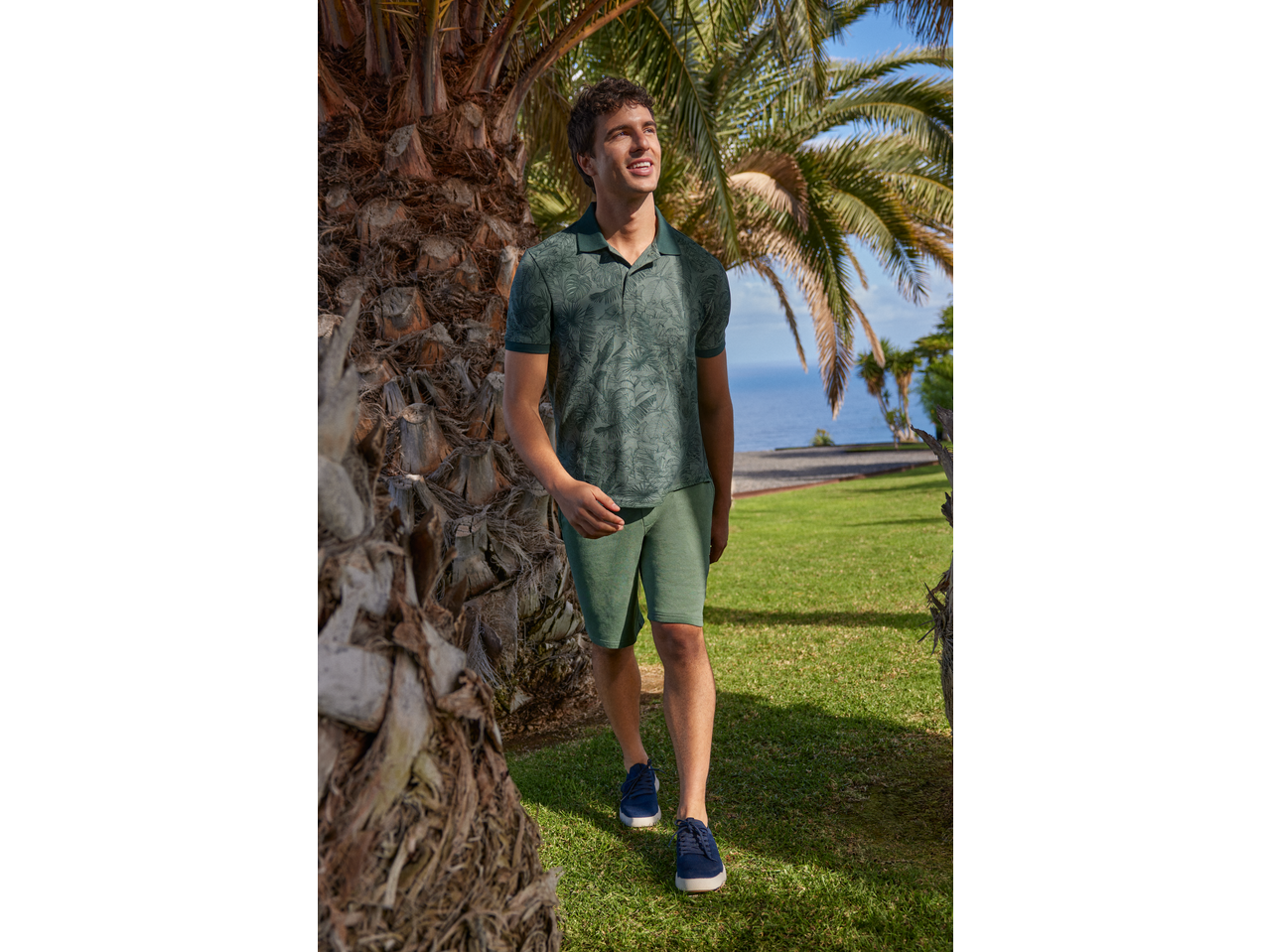 Bărbat în tricou polo verde imprimat și pantaloni scurți verzi, plimbându-se într-o grădină cu palmieri.