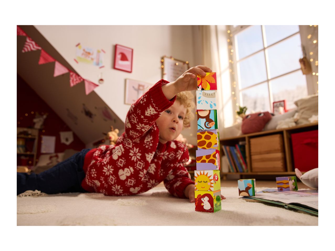 Toddler in a festive jumper playing with stacking blocks on a rug in a decorated room.