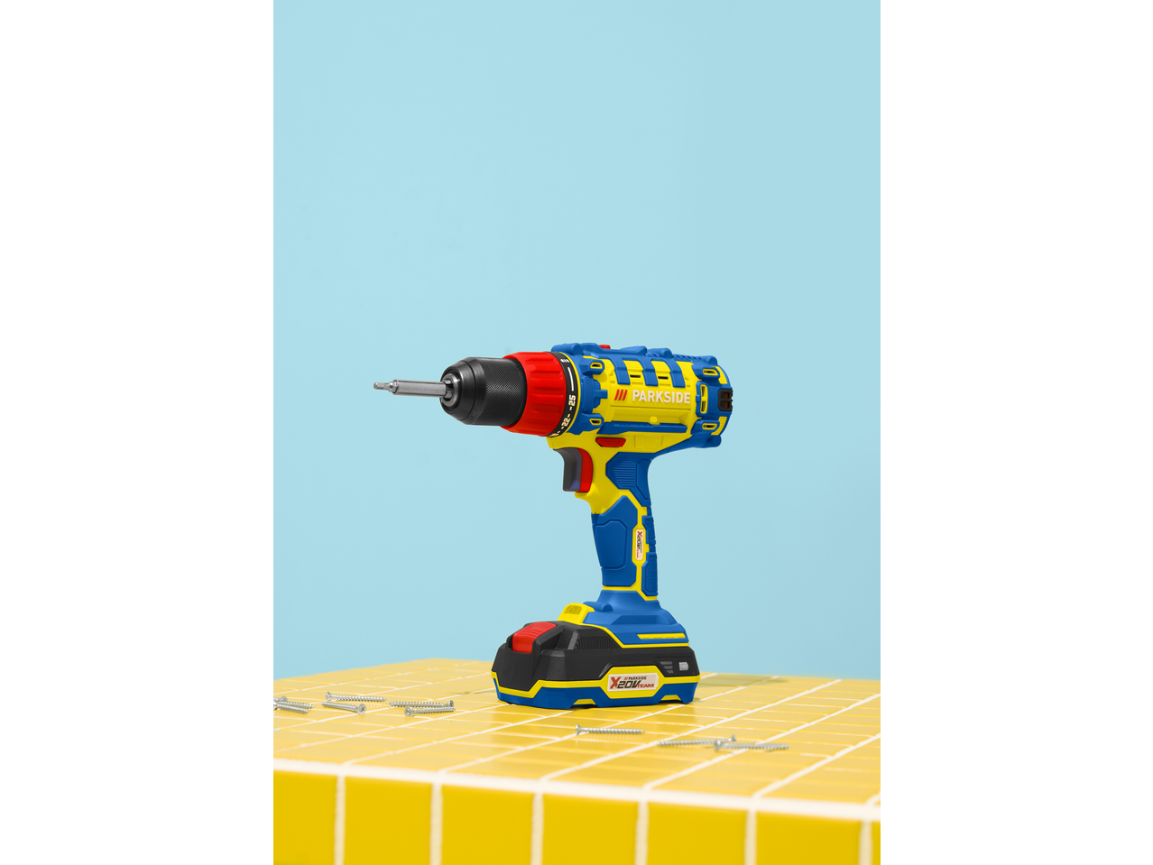 Berbequim sem fios PARKSIDE 20 V azul e amarelo num chão de azulejos amarelos com parafusos.