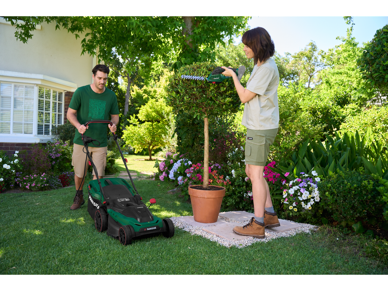 Homem a cortar relva com corta-relvas Parkside®, mulher a podar arbustos com tesoura de podar Parkside®.