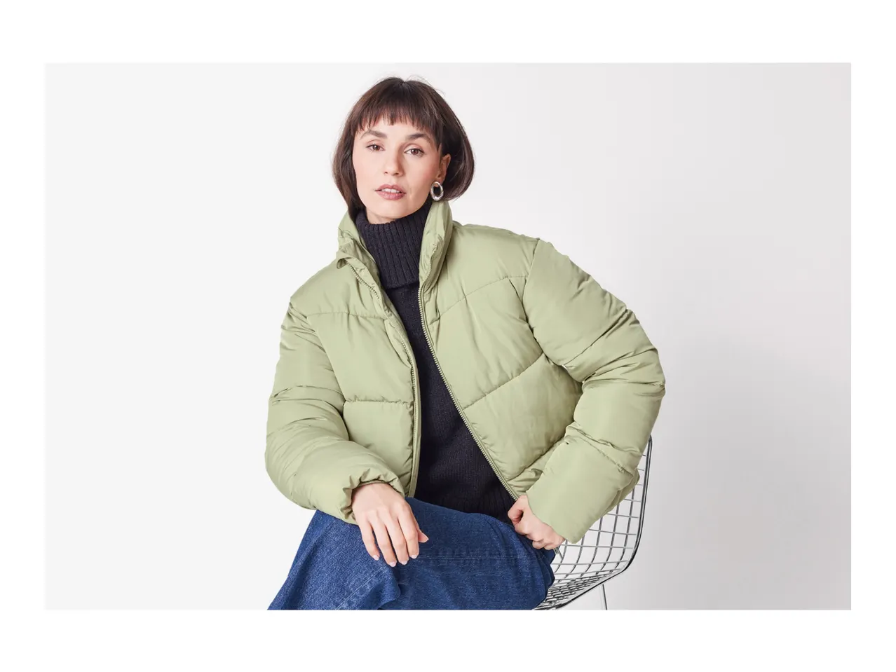 Woman in a light green puffer jacket, black turtleneck, and blue jeans, sitting on a chair.