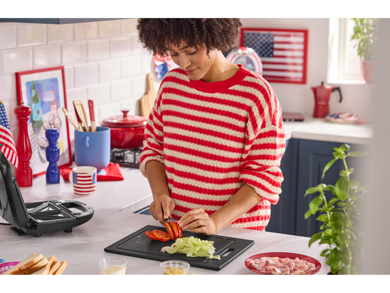 Woman preparing a sandwich with tomato, lettuce, and ham, with a sandwich maker beside her.