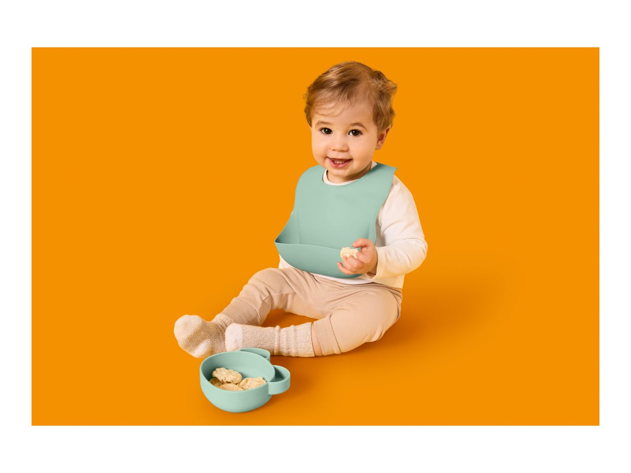 Happy baby in a bib, holding a snack, with a bowl of snacks nearby.