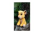 A plush toy of a lion cub with large eyes, sitting on a tree branch amidst green foliage.