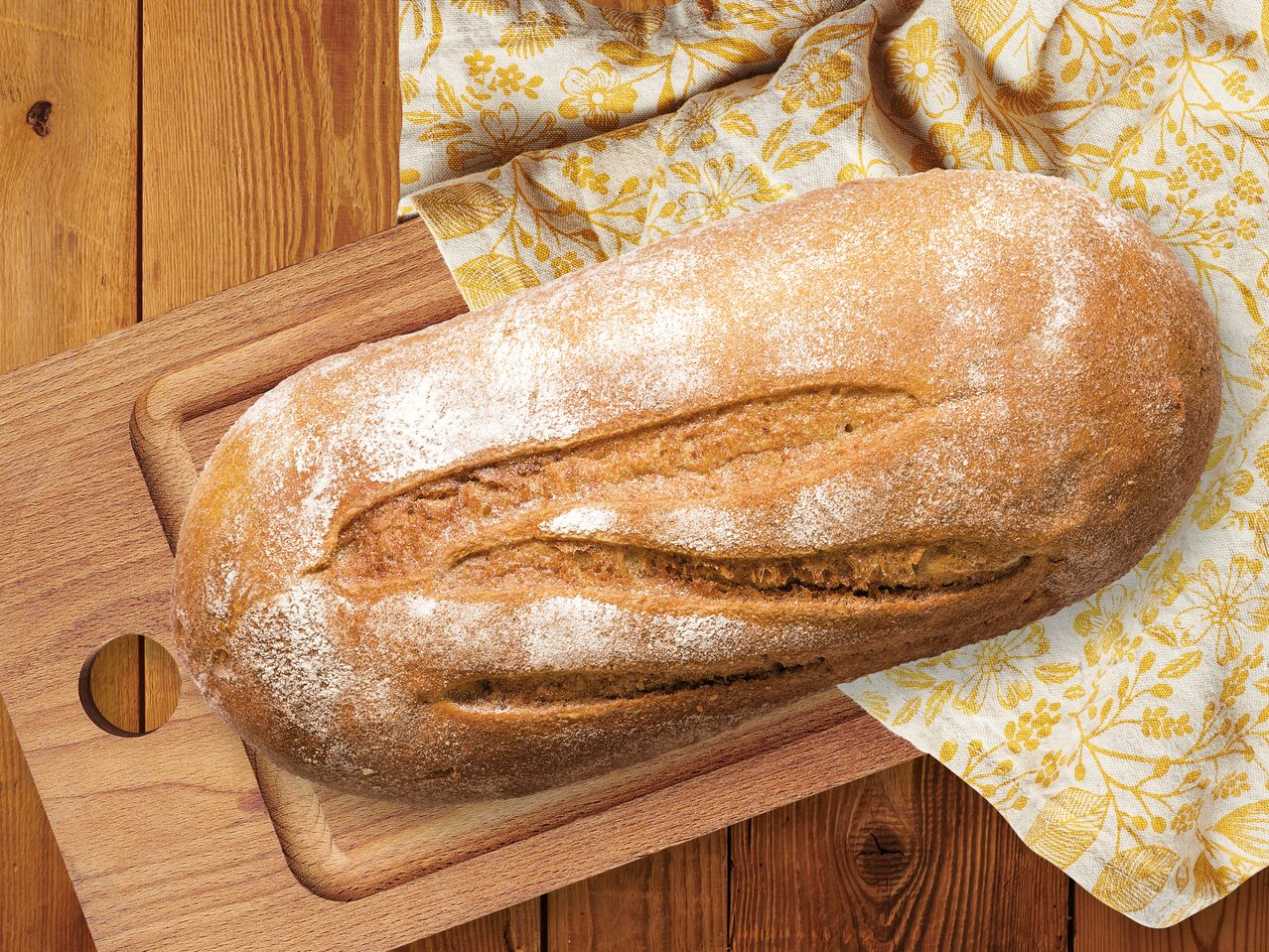 Прясно изпечен хляб върху дървена дъска за рязане и жълта кърпа на дървена маса.