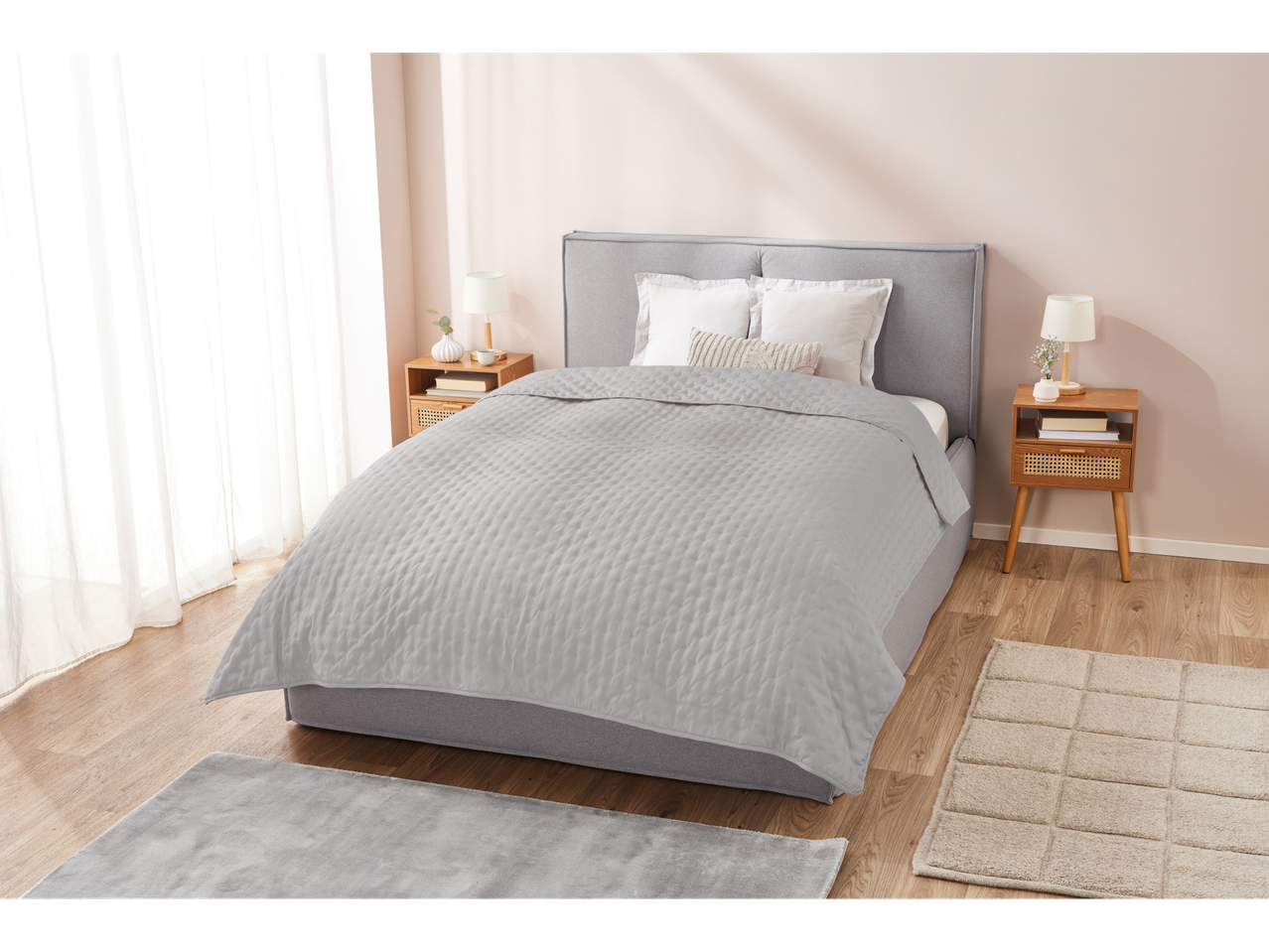A light grey quilted bedspread on a grey upholstered bed in a bright bedroom.