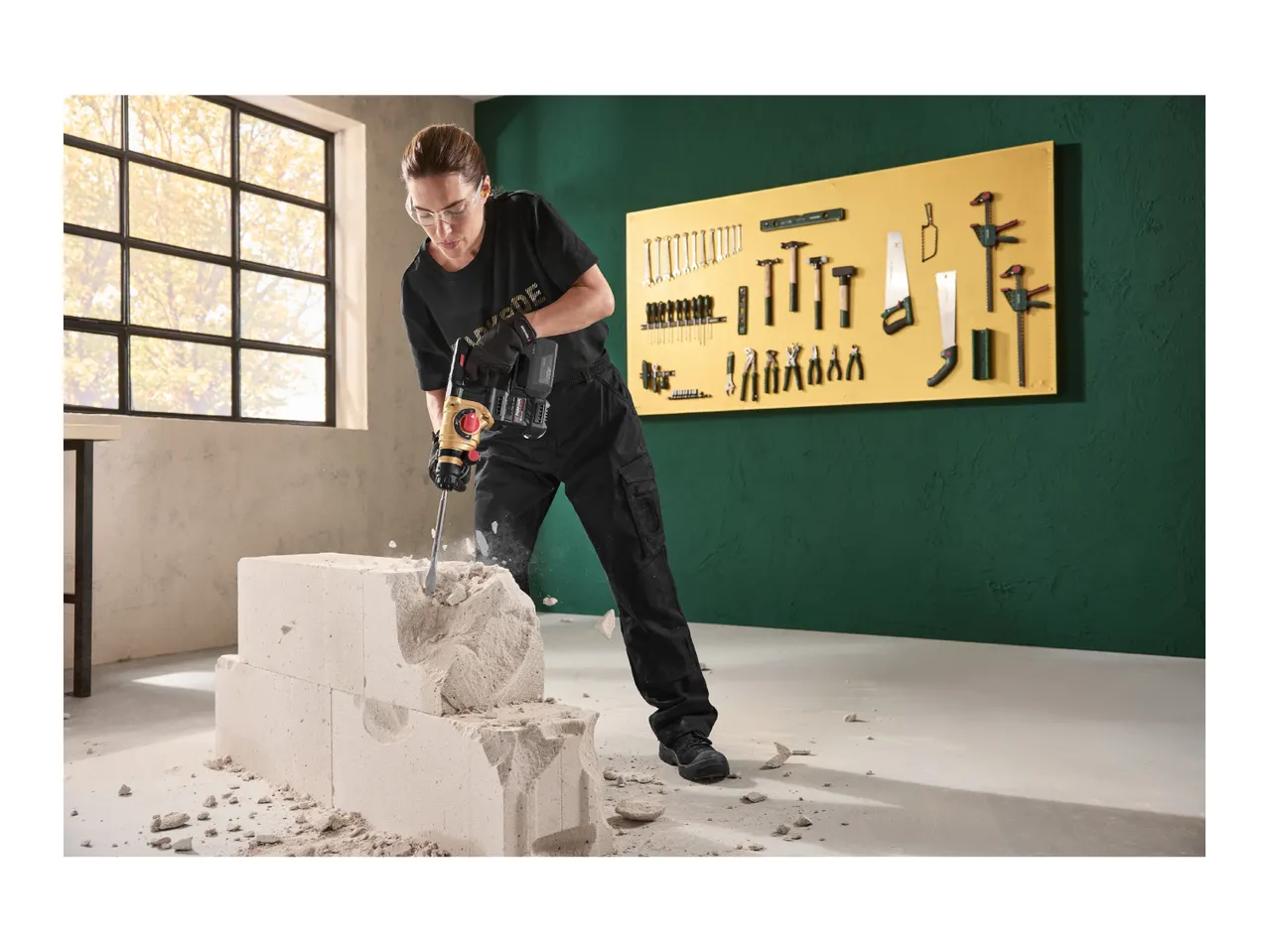 Woman in safety glasses and gloves using a hammer drill on a concrete block.