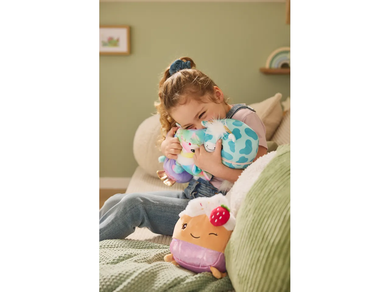 Happy child hugging two plush toys, with another plush toy nearby on a green blanket.