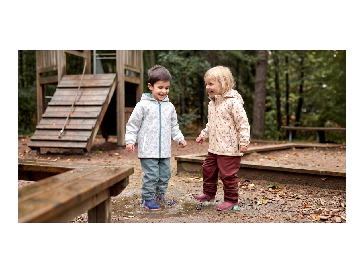 Two happy children in waterproof jackets and trousers splashing in a puddle.