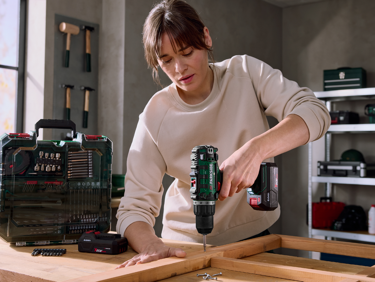 Жена използва акумулаторна бормашина, докато сглобява дървена конструкция, до нея е комплект инструменти.