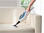 A person uses a handheld steam cleaner to clean a white fabric sofa.