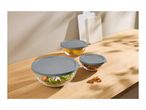 Three glass food storage bowls with grey lids, containing salad, lemons, and pretzels, on a wooden surface.