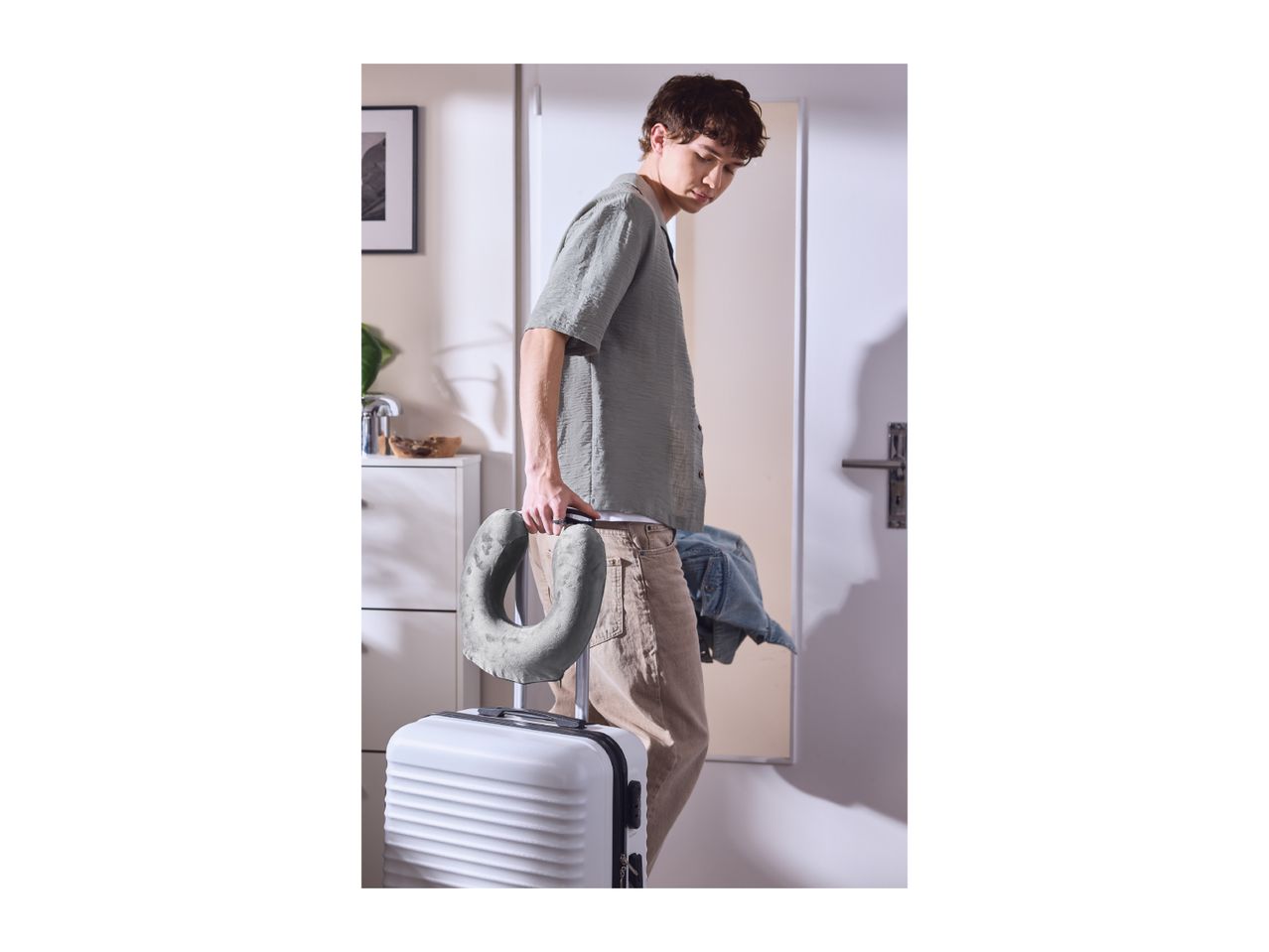 Man with a neck pillow attached to a white suitcase, ready for travel.