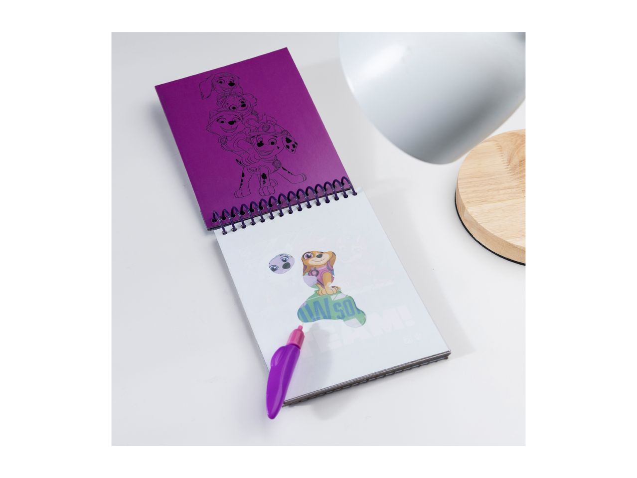 A purple Paw Patrol spiral notebook with a coloring page of Skye and a purple pen on a white surface.