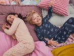 Zwei Kinder in Pyjamas liegen lachend auf einem Bett mit Kissen und Decken.