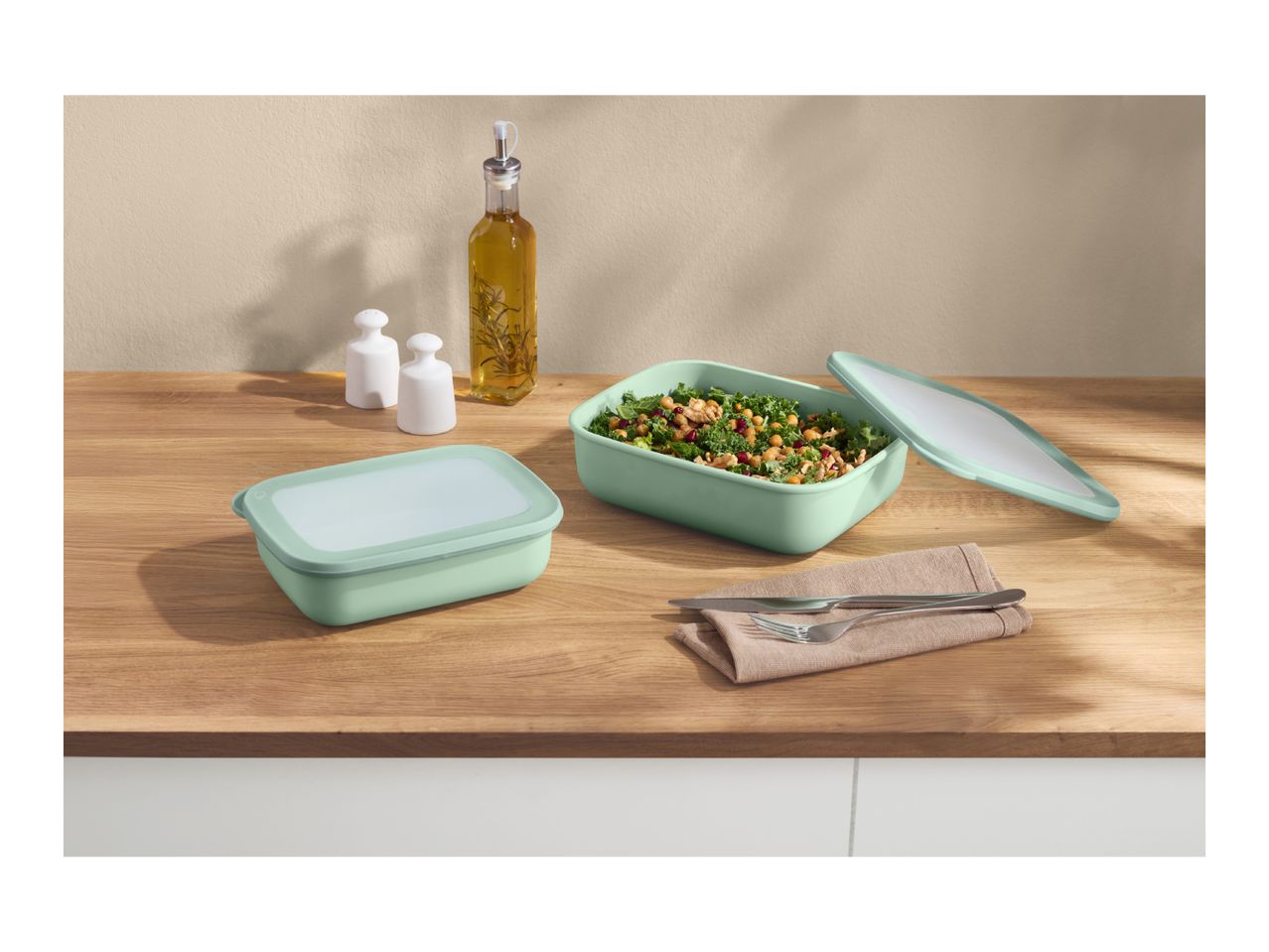 Two light green food containers, one filled with salad, on a wooden counter with olive oil and shakers.