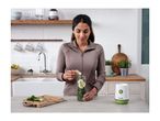 Woman adding cucumber to a blender bottle with greens, next to a personal blender.