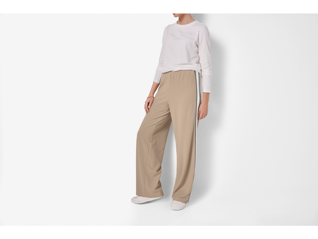 Vrouw in beige trainingsbroek met witte streep en een wit shirt met lange mouwen.