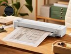 Beli laminator na leseni mizi, ki laminira načrt, z rastlino in pisarniškimi predmeti v ozadju.