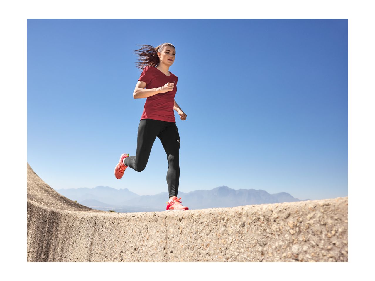 Woman in red t-shirt and black leggings running outdoors with mountains in background.