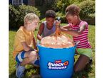Three children filling water balloons from a blue bucket in a sunny garden.