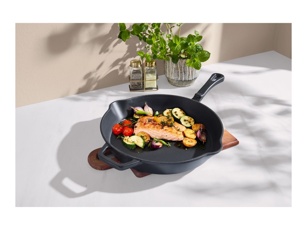 Cast iron pan with salmon, vegetables, and herbs on a white marble surface.