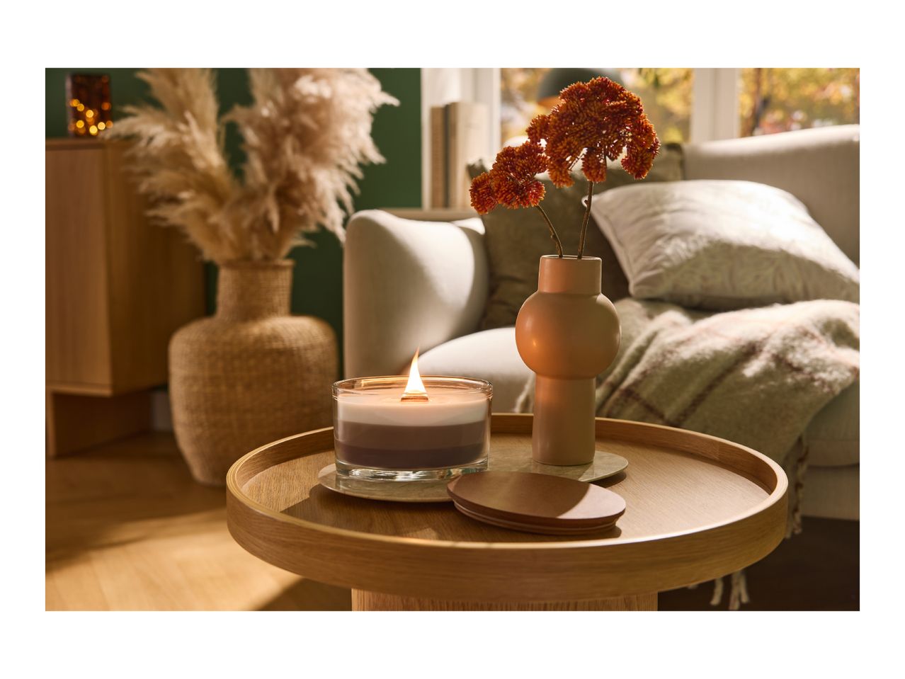 A lit candle and a vase with orange flowers on a wooden side table in a cozy living room.