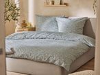 A bedroom with a bed featuring light green floral duvet covers and pillows, a wooden nightstand, and a plant.