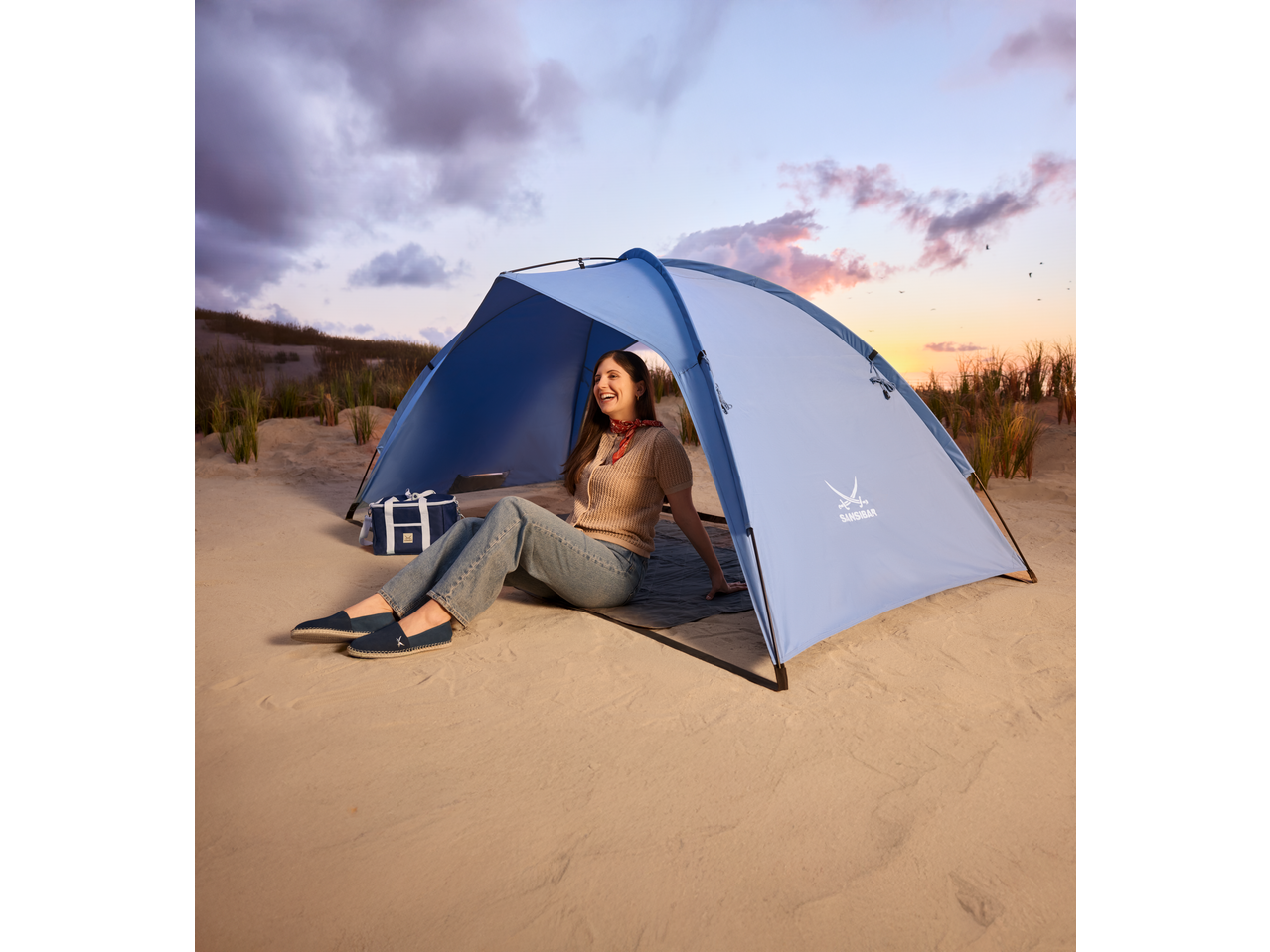 Lächelnde Frau in einer blauen SANSIBAR Strandmuschel an einem Sandstrand bei Sonnenuntergang.
