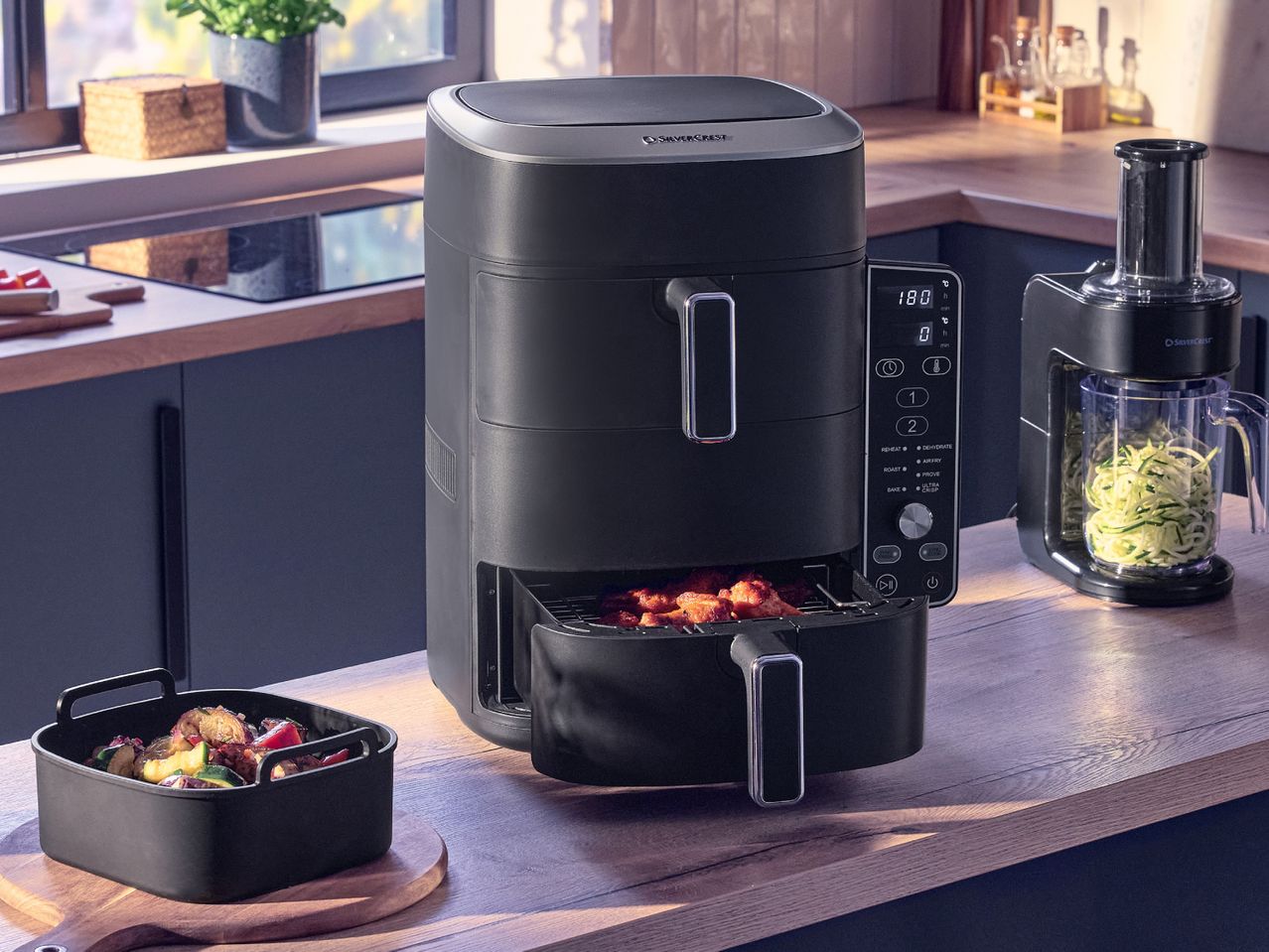 Black air fryer with food inside, digital display showing 180 degrees, and a spiralizer with zucchini noodles on a kitchen counter.