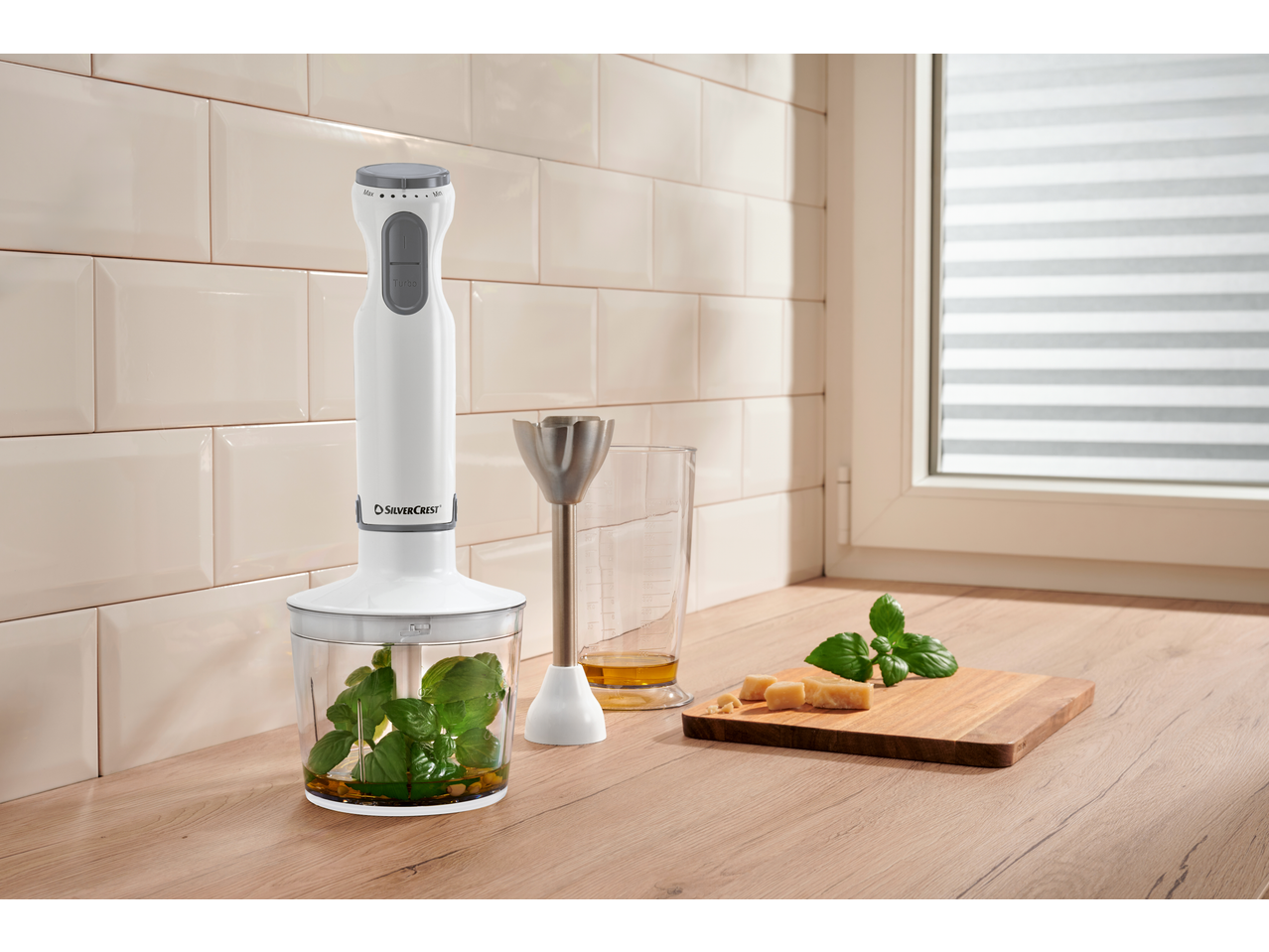 Hand blender with basil and oil in a chopper, next to a measuring cup and cheese.