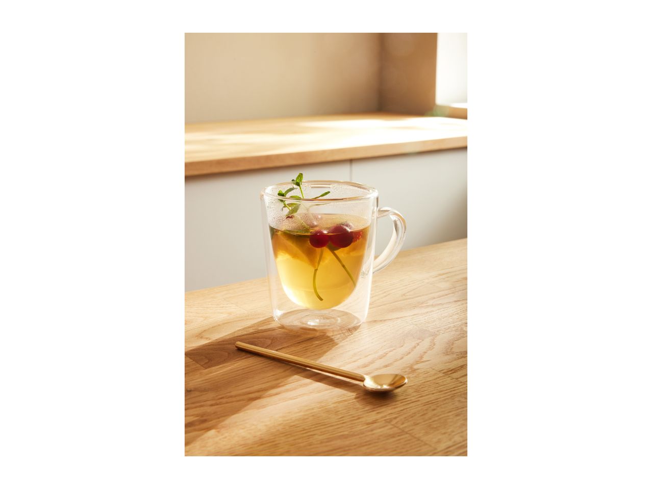 Glass mug with tea, cherries, and mint on a wooden table with a golden spoon.