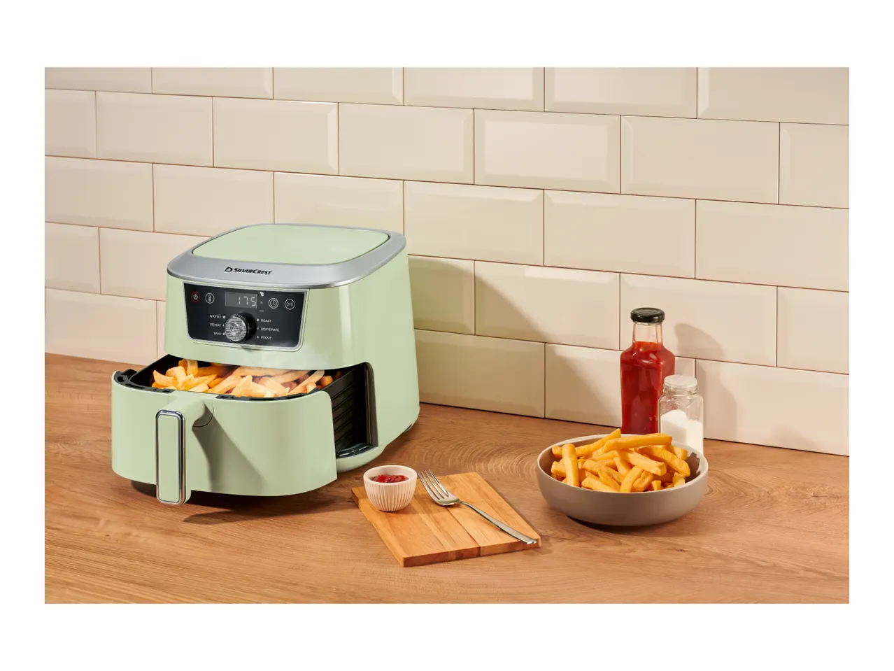 Green air fryer with fries, ketchup, and salt on a wooden counter.