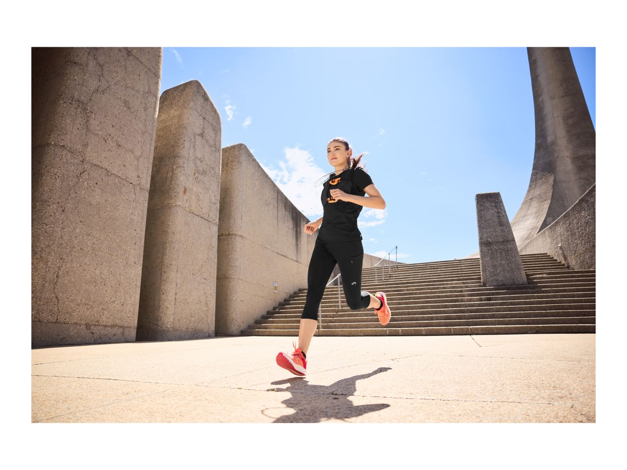 Woman running in black activewear and orange running shoes on concrete steps.